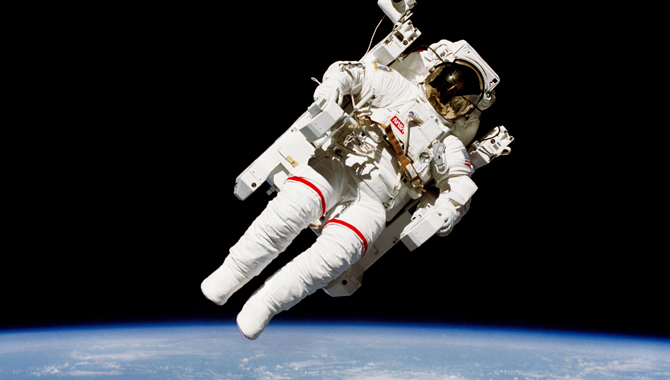 Astronauta w białym skafandrze kosmicznym unoszący się nad Ziemią podczas spaceru kosmicznego, z widoczną krzywizną planety w tle.